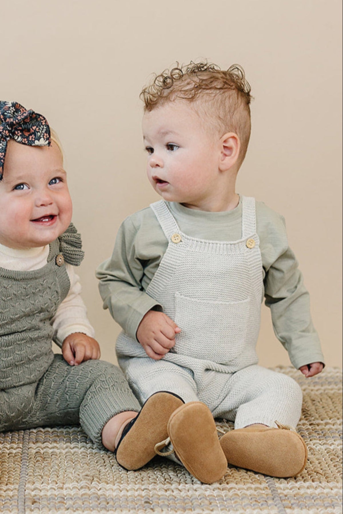 Two toddlers sitting on a woven rug, wearing knit overalls and sweaters, with one wearing a floral headband.