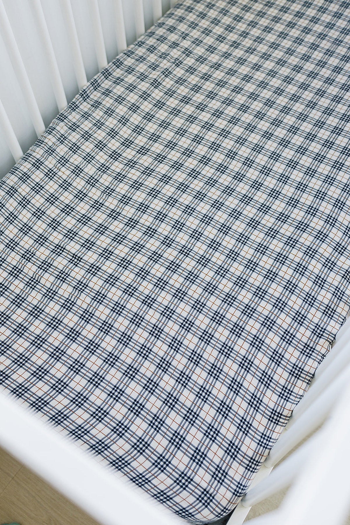 A white crib features a vintage plaid muslin crib sheet with blue and brown lines, made from 100% cotton.