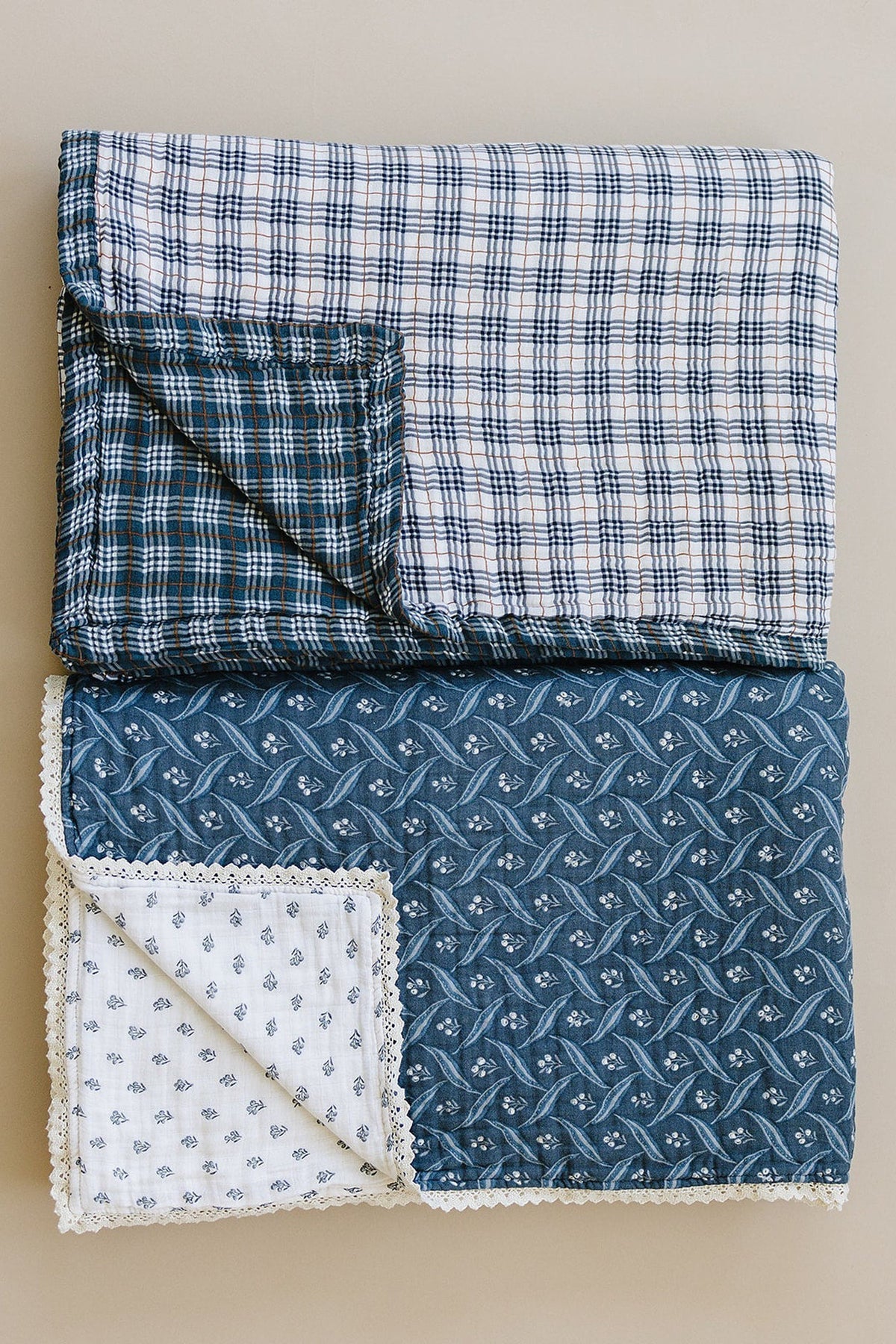 Folded quilt with blue plaid and floral patterns, featuring lace edges on a beige background. Made of cotton.
