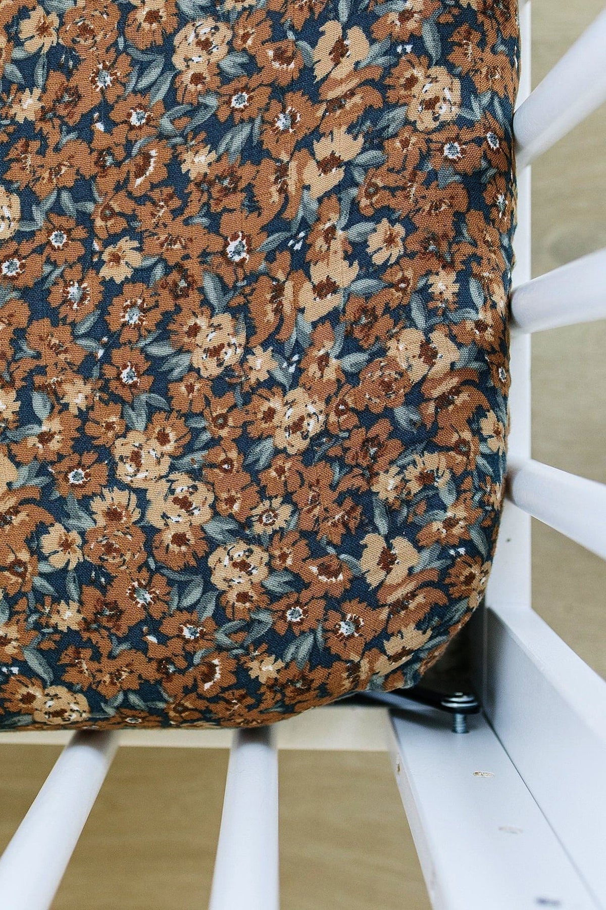 Floral-patterned muslin crib sheet with brown and blue flowers, fitted on a white crib. Made from soft cotton.