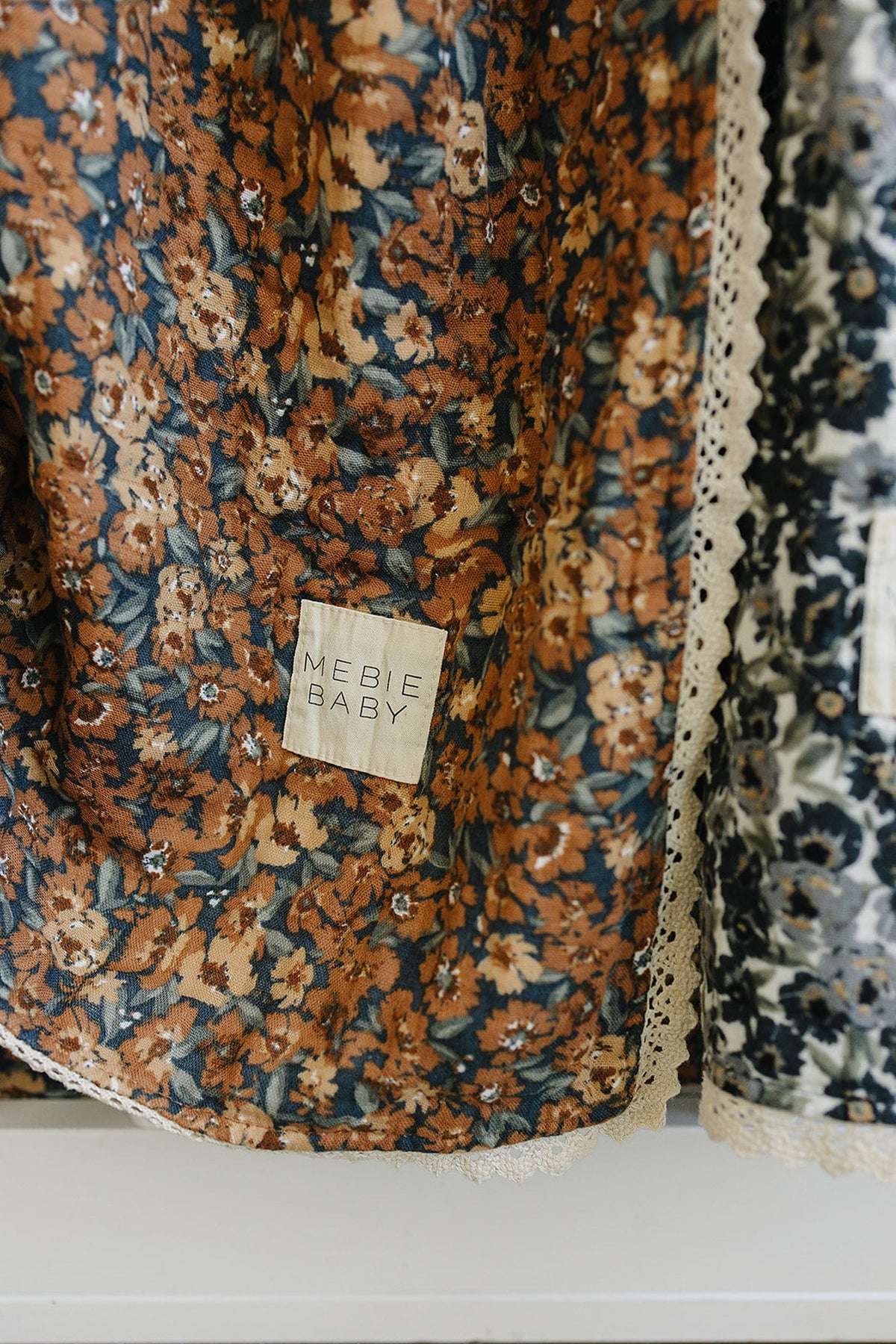 Floral quilt with lace edges, featuring a Mebie Baby label, in warm brown and navy tones.