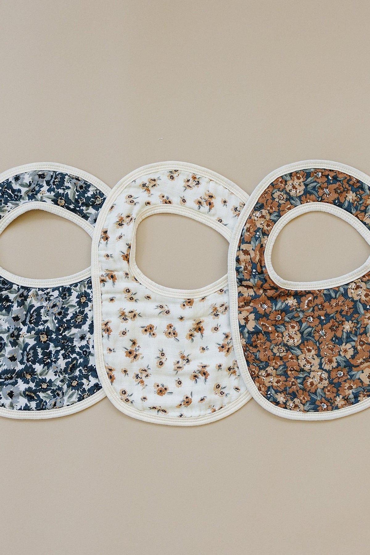 Three floral-patterned muslin bibs in blue, white, and brown on a beige background, made from 100% cotton.