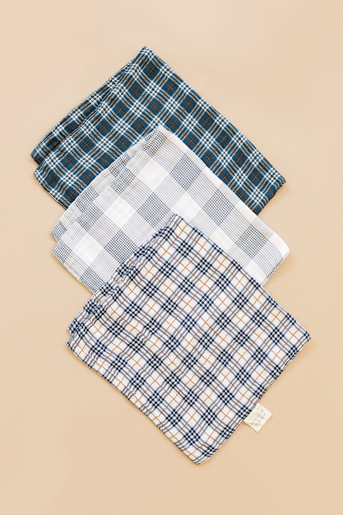 Three muslin burp cloths featuring navy and white plaid patterns are stacked on a neutral background. Each cloth is soft, made from 100% cotton, ideal for baby essentials.