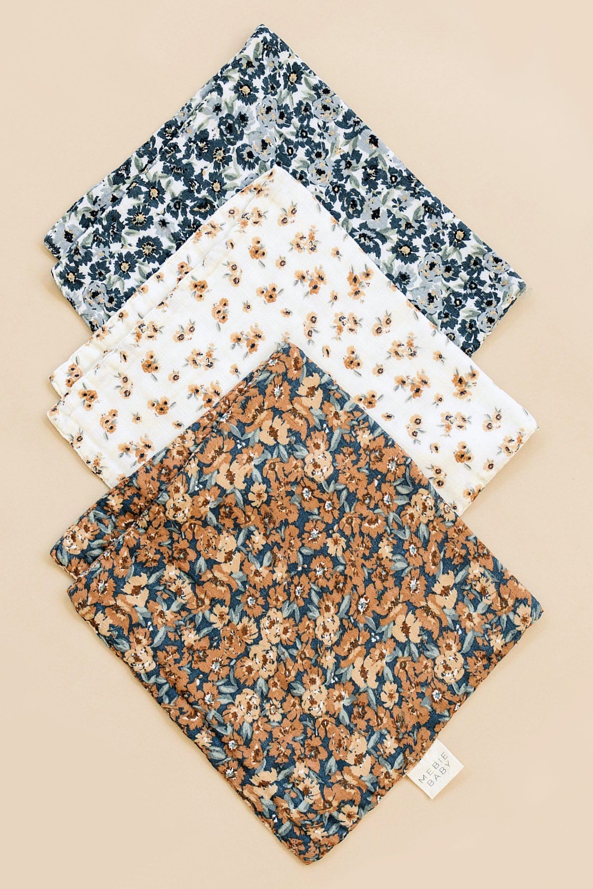 Three floral muslin burp cloths in blue, white, and brown patterns are laid on a beige background.