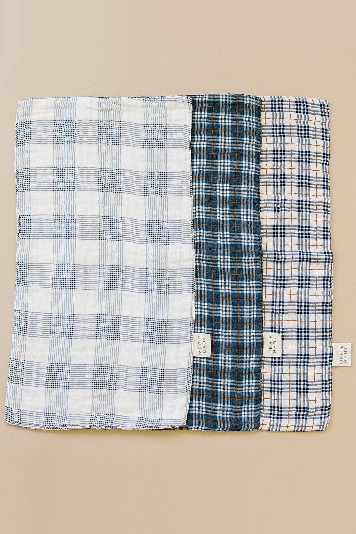 Three Mebie Baby Coastal Plaid Muslin Burp Cloths in blue, beige, and navy plaid patterns on a beige background.