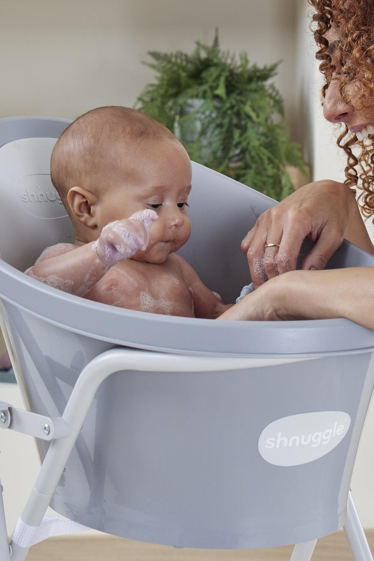A baby is bathed in a gray Shnuggle bathtub, supported by an adults hand.