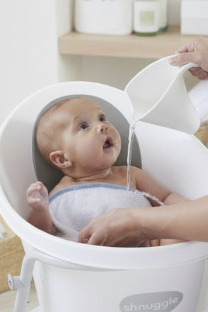 A baby in a white Shnuggle bath is being rinsed with water from a white jug.