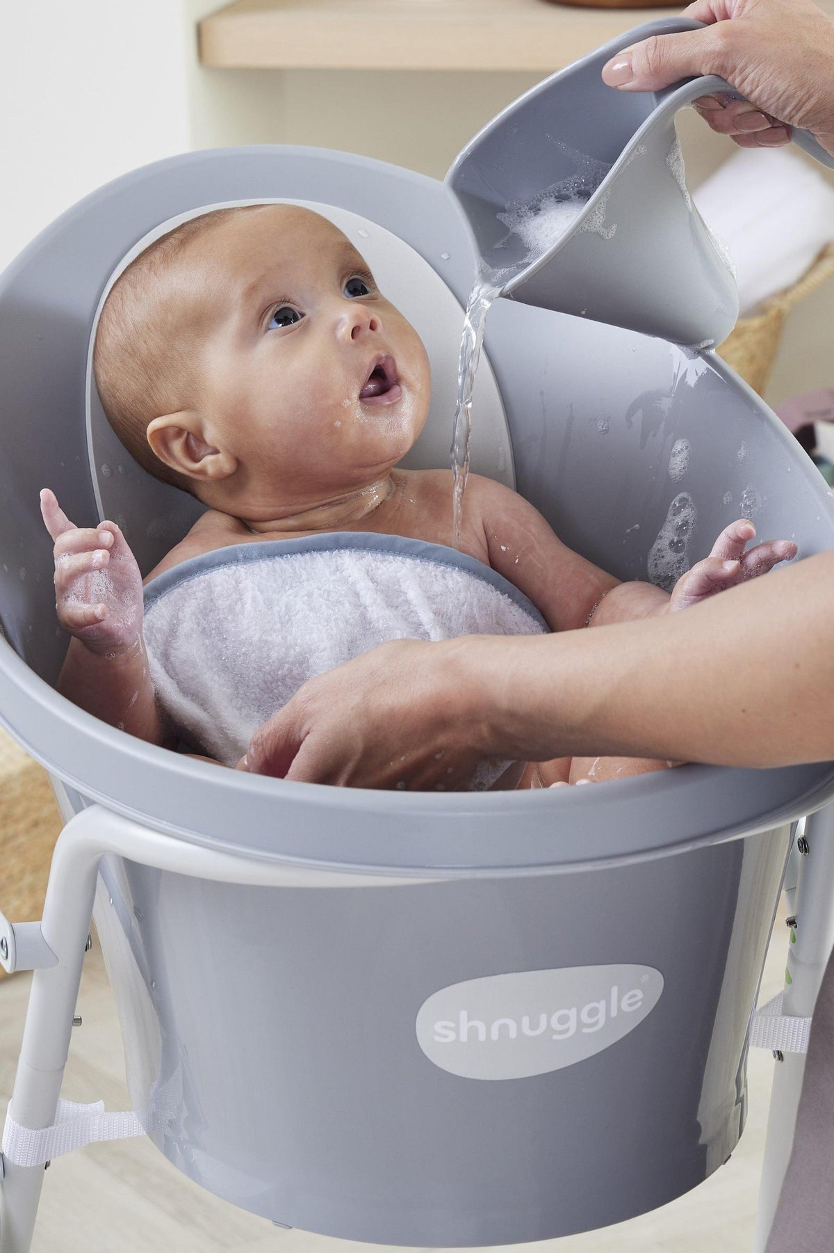 A baby is bathed in a Shnuggle tub while an adult pours water from a gray jug.