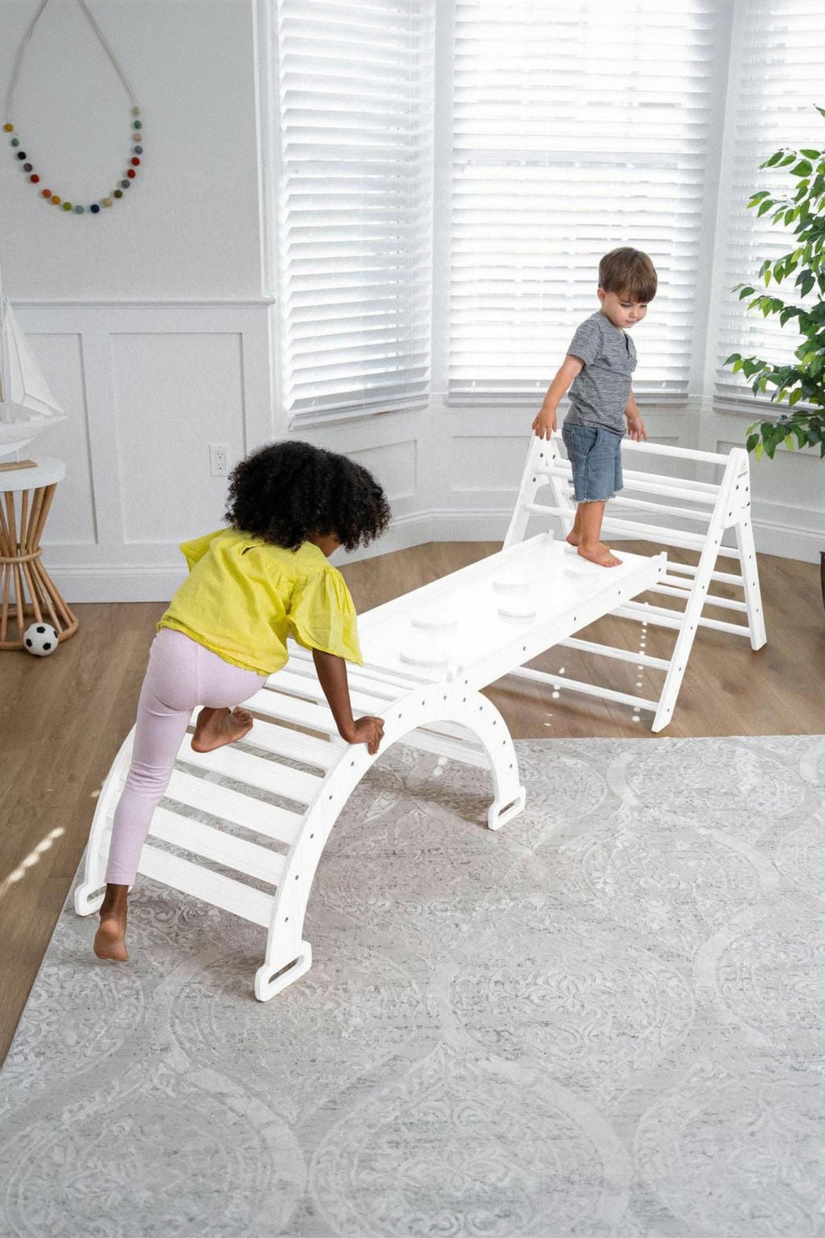 Two children play on a white wooden Pikler triangle playset inside a bright room with large windows. One child climbs while the other balances on the top, showcasing active play.