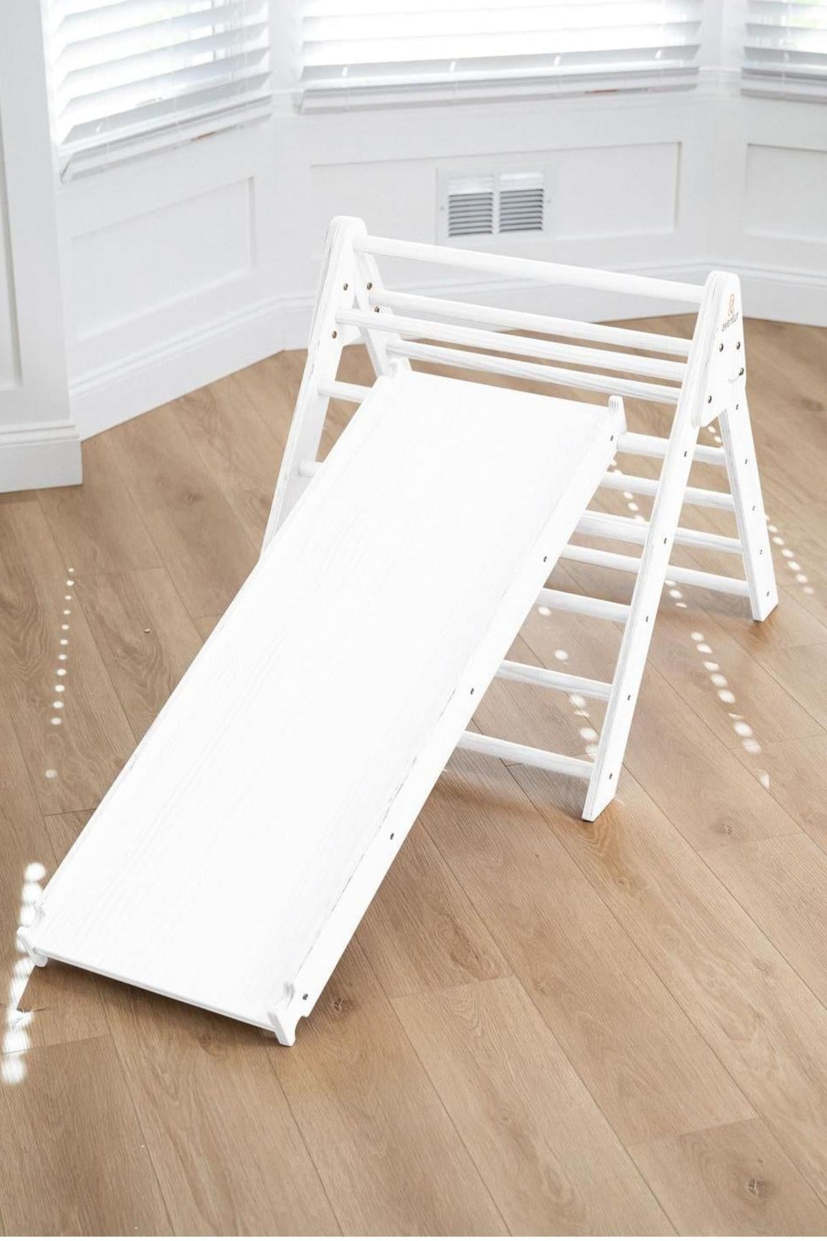 A white wooden Pikler triangle with a slide attached, set on a light wooden floor in a bright room with natural light. Ideal for active play and safe indoor adventures.