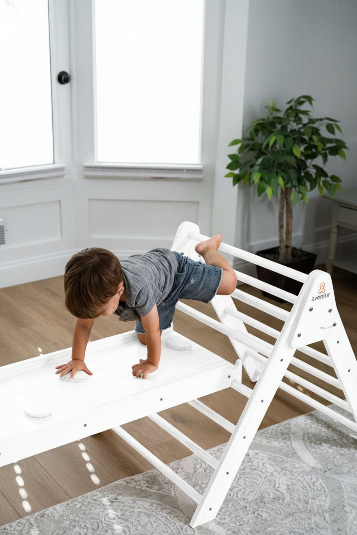 A young child in a gray shirt climbs on a white wooden Pikler triangle playset indoors, showcasing active play. Sunlight filters through the windows, illuminating the space.