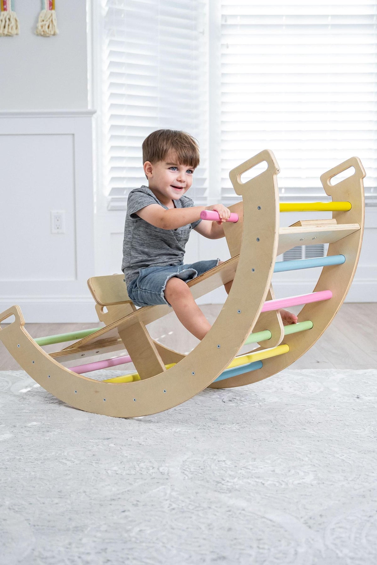 A young child joyfully plays on a wooden Bailis climbing arch that doubles as a rocker, featuring colorful grips. The bright, airy room has white walls and natural light streaming.
