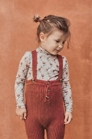 A toddler stands against a textured orange wall, wearing cozy, ribbed rosewood knit overalls with button details and a floral long-sleeve shirt, complemented by brown shoes.