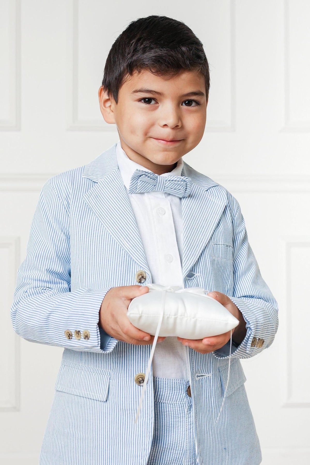 A young boy smiles while holding a small, white cushion. He wears a light blue seersucker suit jacket with a bow tie, showcasing a classic.