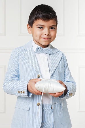 A young boy smiles while holding a small, white cushion. He wears a light blue seersucker suit jacket with a bow tie, showcasing a classic.