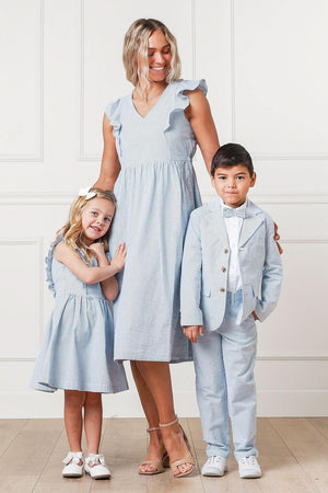 A woman and two children pose together, all wearing light blue outfits. The boy is in a tailored suit with a bow tie, while the girl wears a matching dress.