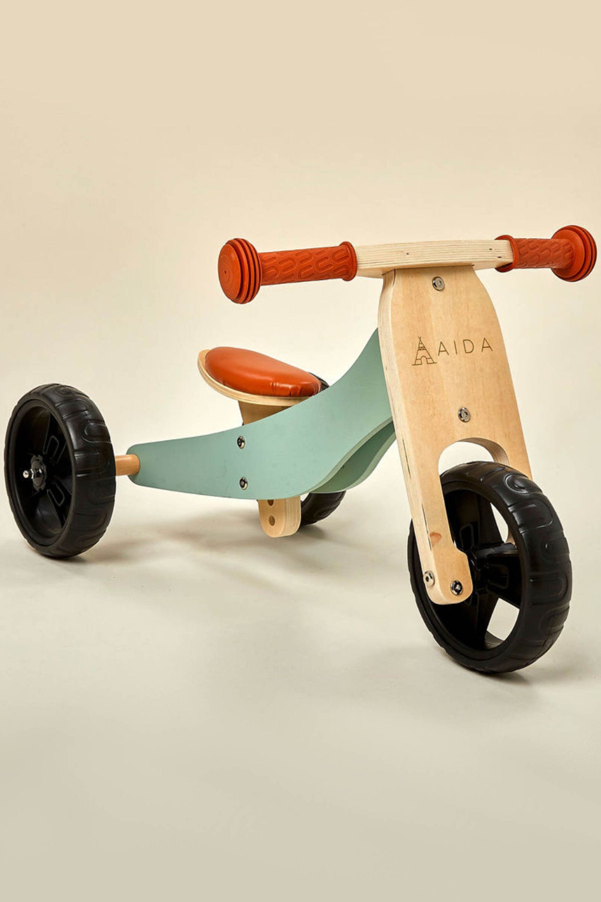 A wooden balance bike featuring a green seat and handlebar grips, designed for toddlers aged 18 months to 3 years.