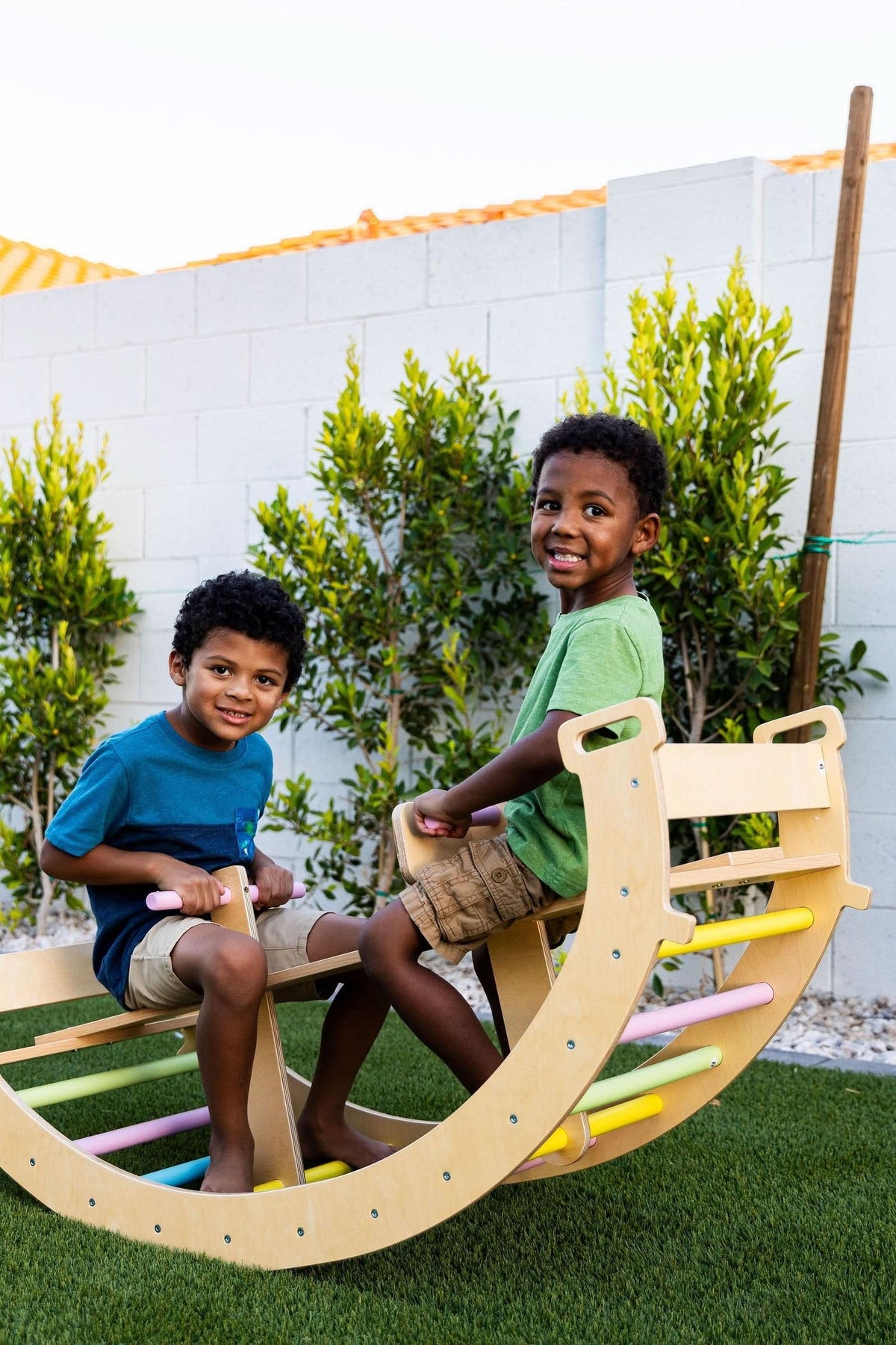 Two young boys play joyfully on a wooden climbing arch, which features a natural finish and colorful rungs, set against a backdrop of neatly trimmed greenery and a white wall.