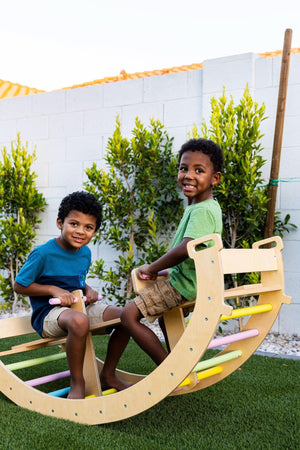 Two young boys play joyfully on a wooden climbing arch, which features a natural finish and colorful rungs, set against a backdrop of neatly trimmed greenery and a white wall.