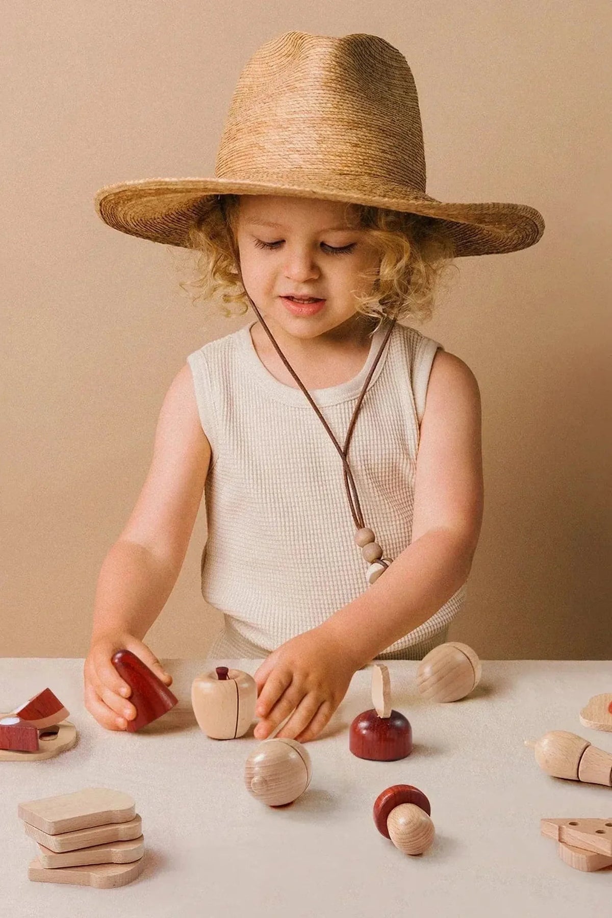 A young child wearing a wide-brimmed straw hat engages with a colorful wooden food set, arranging various food pieces on a light-colored surface against a neutral backdrop.