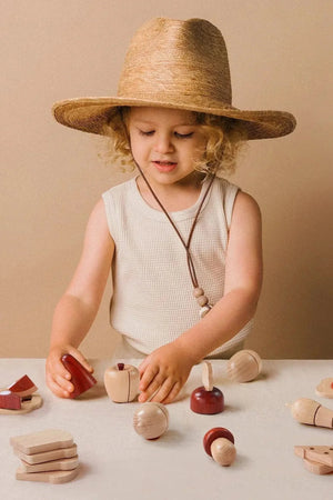 A young child wearing a wide-brimmed straw hat engages with a colorful wooden food set, arranging various food pieces on a light-colored surface against a neutral backdrop.