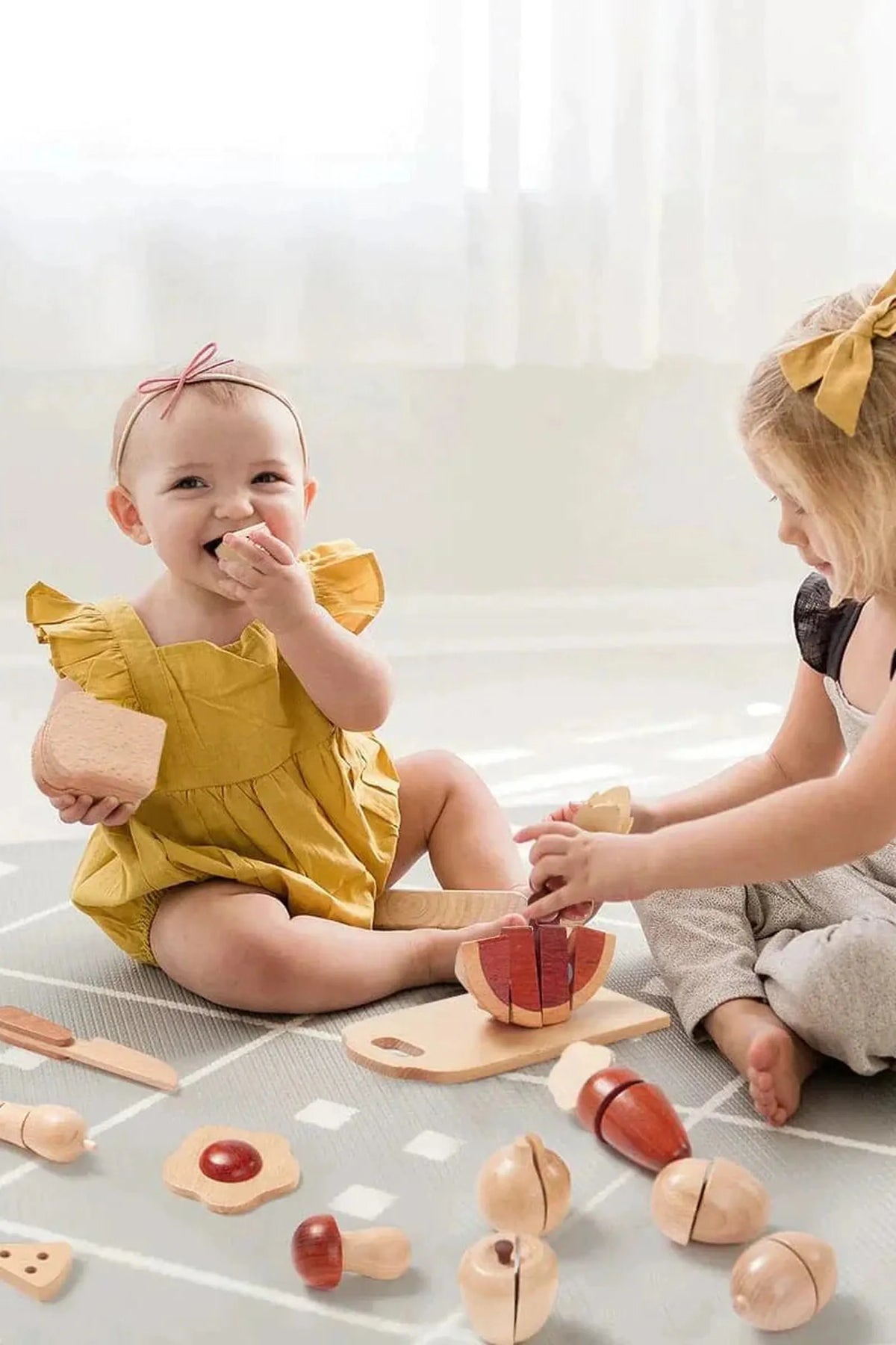 Two young children sit on a soft, patterned play mat, engaged in a wooden food play set.