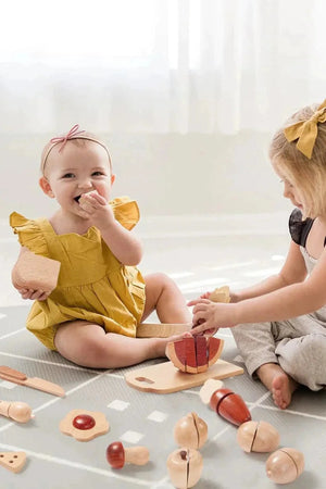 Two young children sit on a soft, patterned play mat, engaged in a wooden food play set.