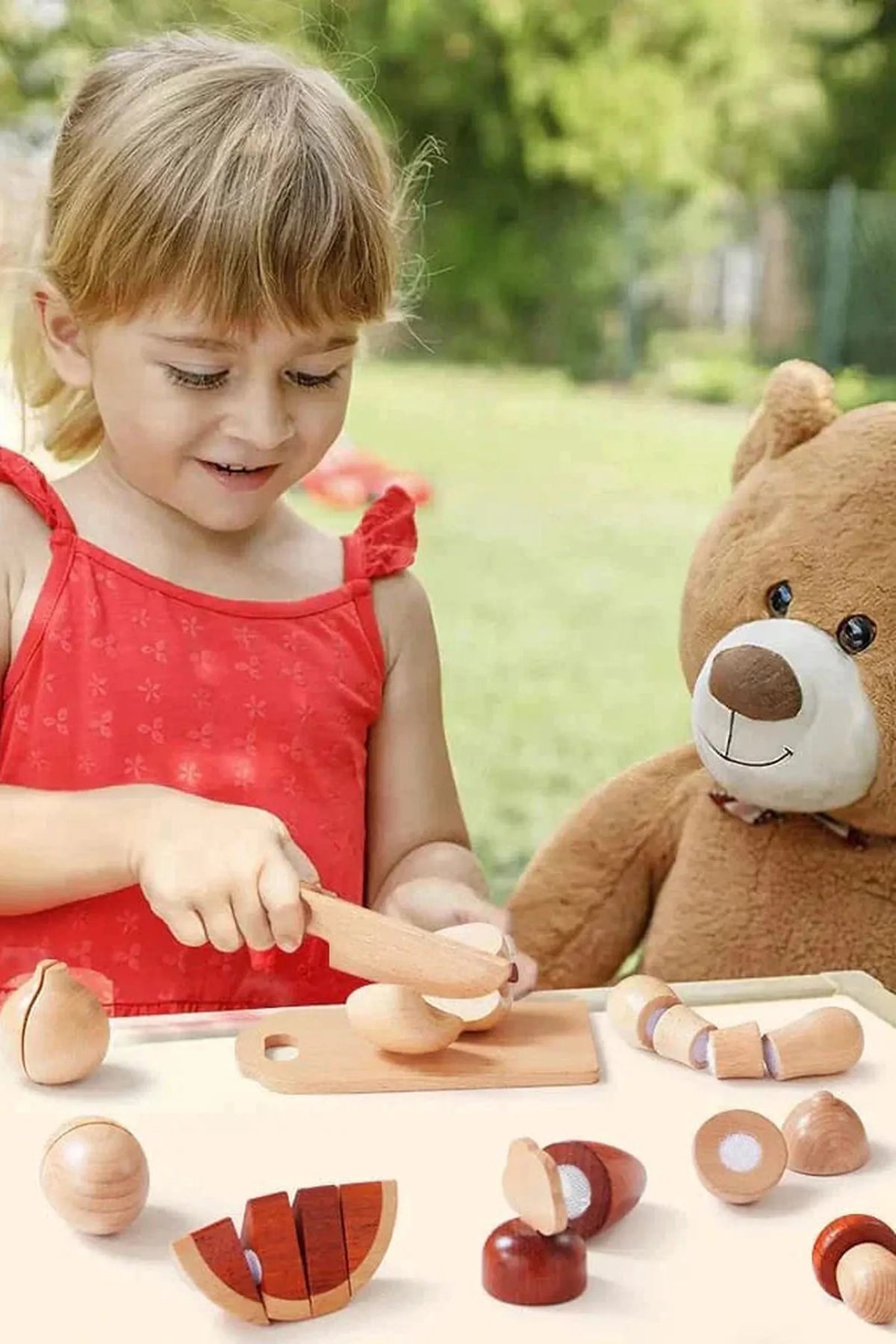 A young girl in a red dress joyfully slices wooden food pieces on a table while a teddy bear sits beside her in a sunny outdoor setting.