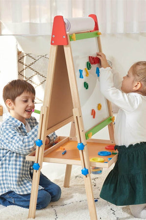 Two children engage with a wooden easel featuring a roll of paper on top, colorful magnetic shapes, and drawing tools.