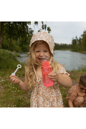 A joyful child with long, blonde hair wears a floral dress and a light-colored bucket sun hat, holding a pink bubble bottle by a tranquil riverside surrounded by greenery.