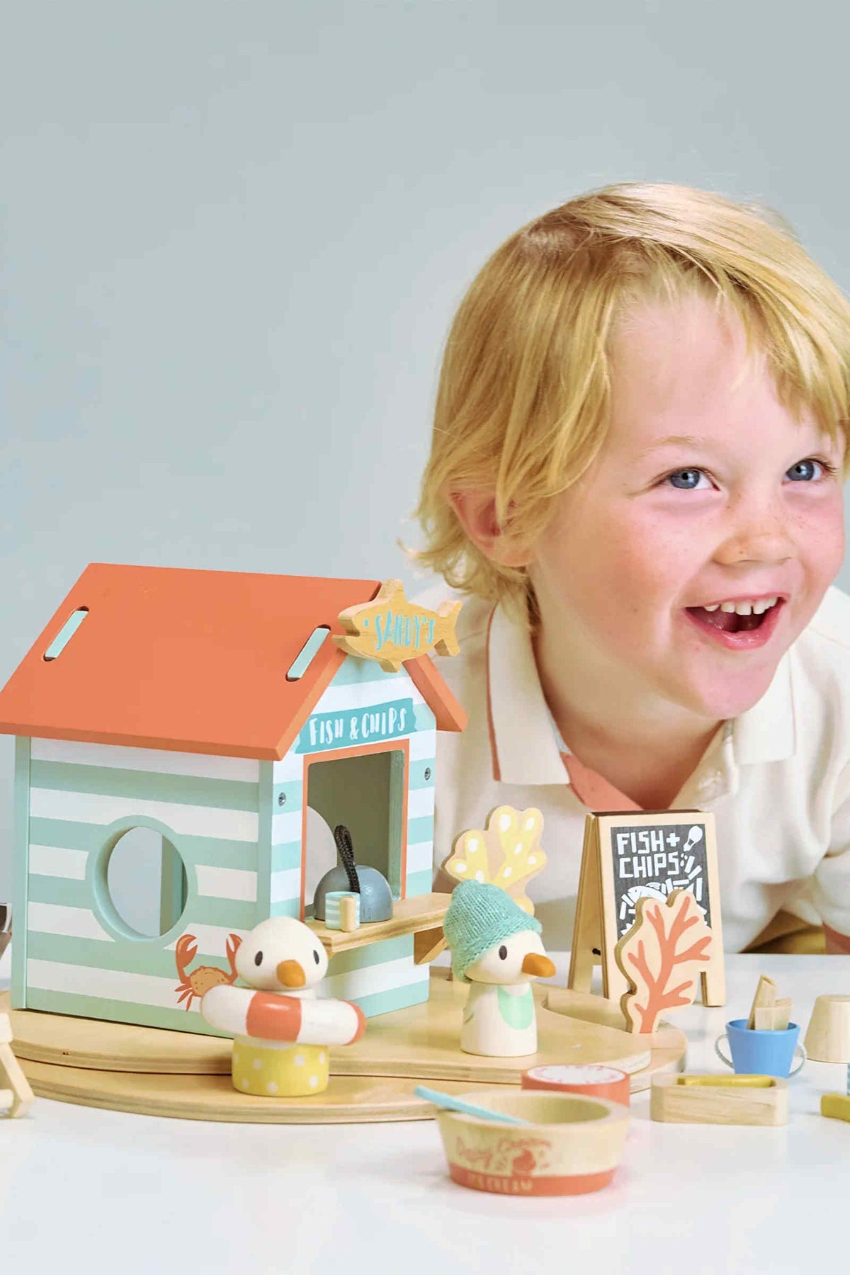A joyful child plays with the Tender Leaf Wooden Toys beach hut set, featuring a colorful wooden seagull and accessories.