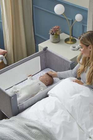 A mother gently touches her sleeping baby in a gray Shnuggle Air Lite Bedside Bassinet, featuring breathable mesh sides.