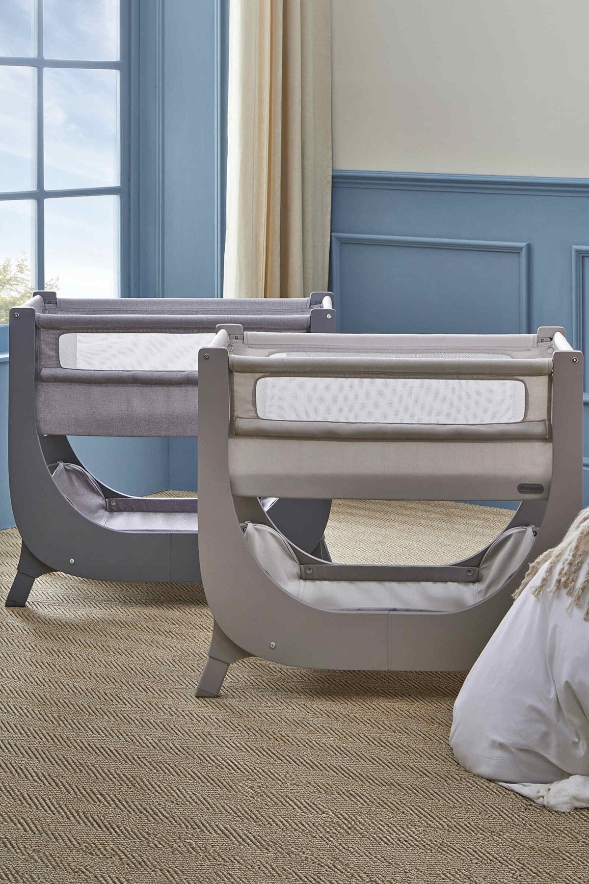 Two Shnuggle Air Lite Bedside Bassinets in soft gray and light gray, featuring breathable mesh sides and a sleek wooden frame, positioned in a bright room with blue walls.