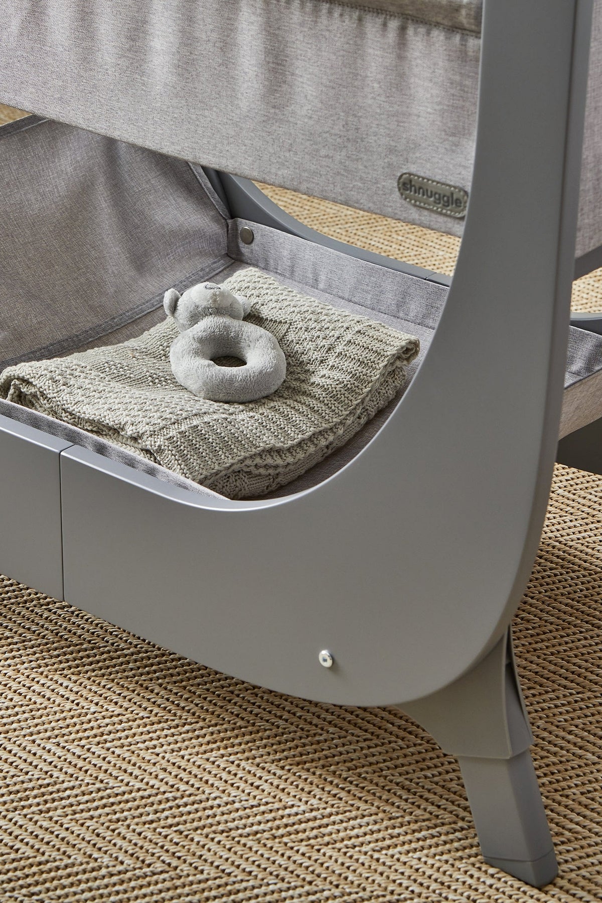 A gray Shnuggle Air Lite Bedside Bassinet features a soft fabric interior with a cozy blanket and a plush toy inside.