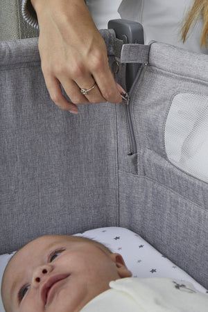 A close-up of a hand adjusting the zipper on the Shnuggle Air Lite Bedside Bassinet, featuring a soft gray fabric and breathable mesh sides, with a baby lying inside.
