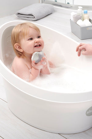 A smiling toddler plays with a toy in a white Shnuggle Toddler Bath filled with bubbles.