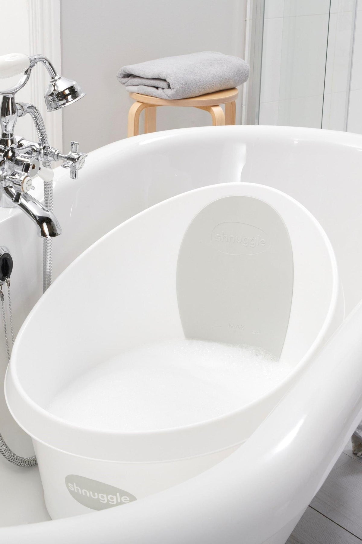 White Shnuggle Toddler Bath Tub with foam, placed in a larger bathtub beside a wooden stool with a towel.