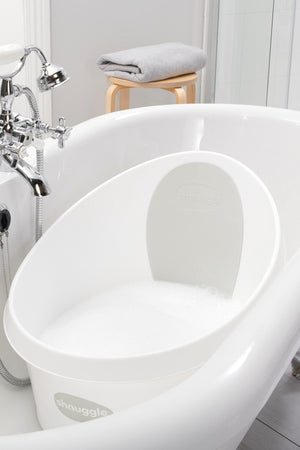 White Shnuggle Toddler Bath Tub with foam, placed in a larger bathtub beside a wooden stool with a towel.