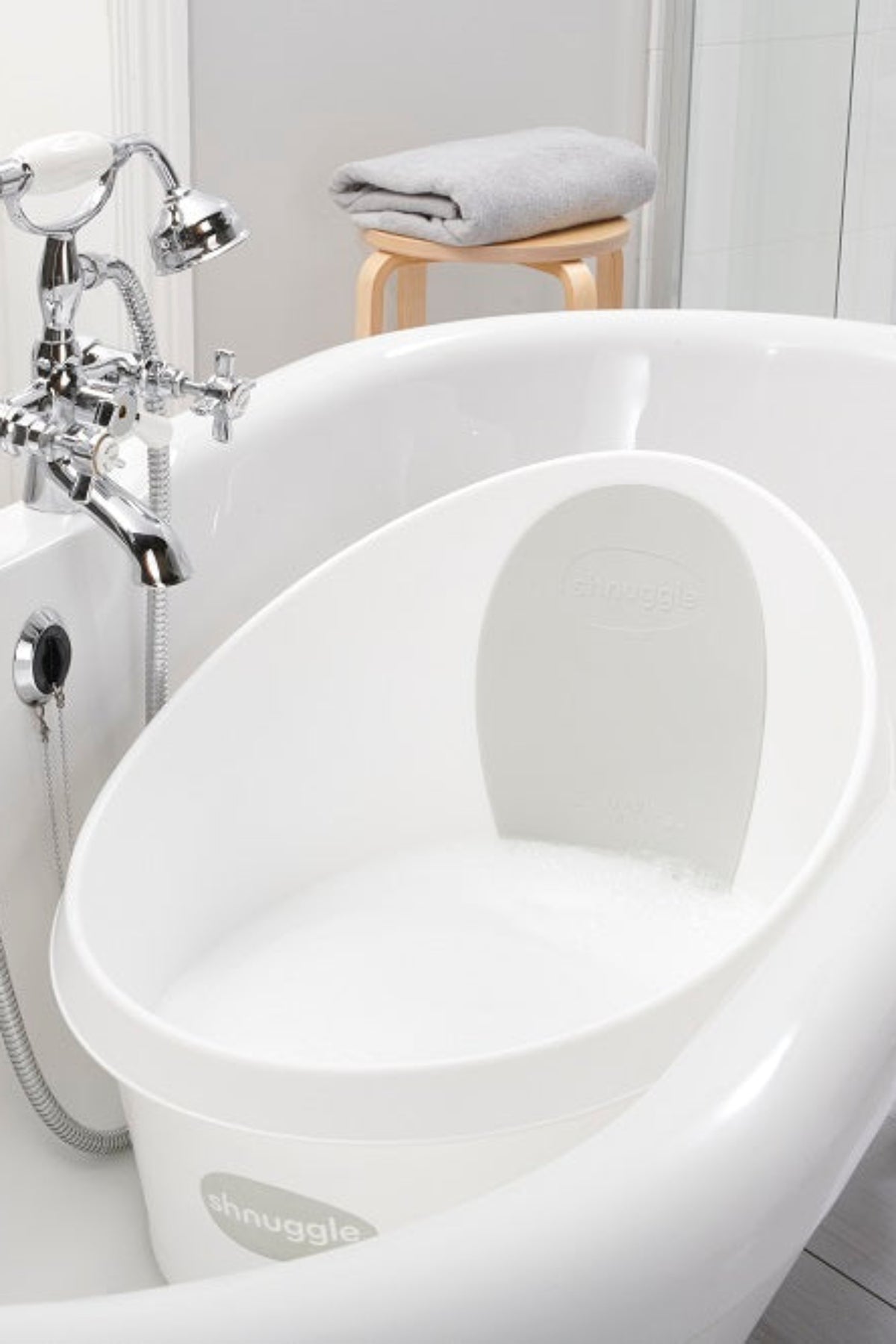 White Shnuggle Toddler Bath Tub inside a larger bathtub, with chrome faucet and a folded towel on a stool.