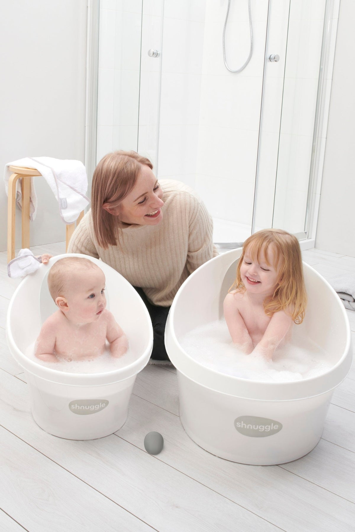 A woman smiles at two children in white Shnuggle bath tubs filled with bubbles in a bright bathroom.