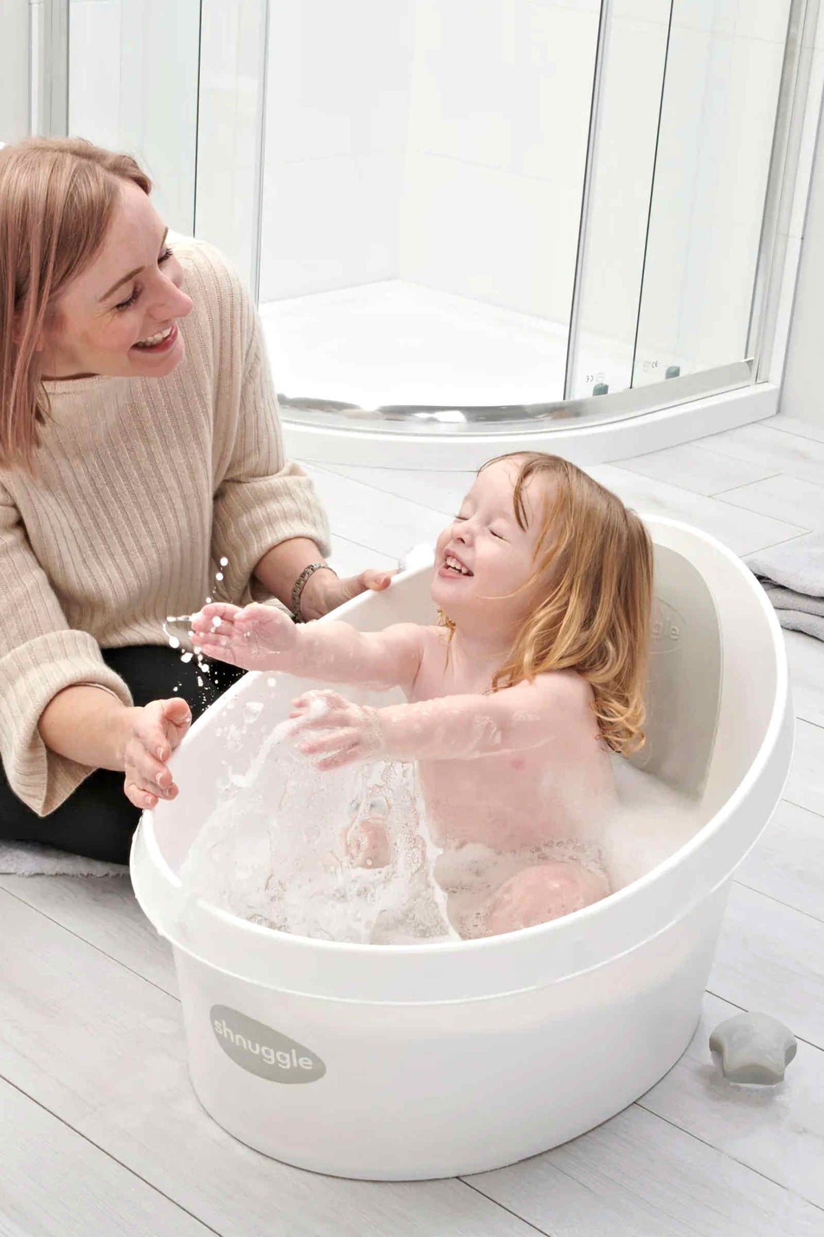 A happy toddler splashes in a white Shnuggle bath tub, with an adult smiling nearby in a bright bathroom.