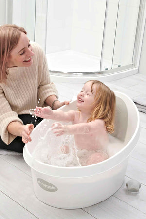 A happy toddler splashes in a white Shnuggle bath tub, with an adult smiling nearby in a bright bathroom.