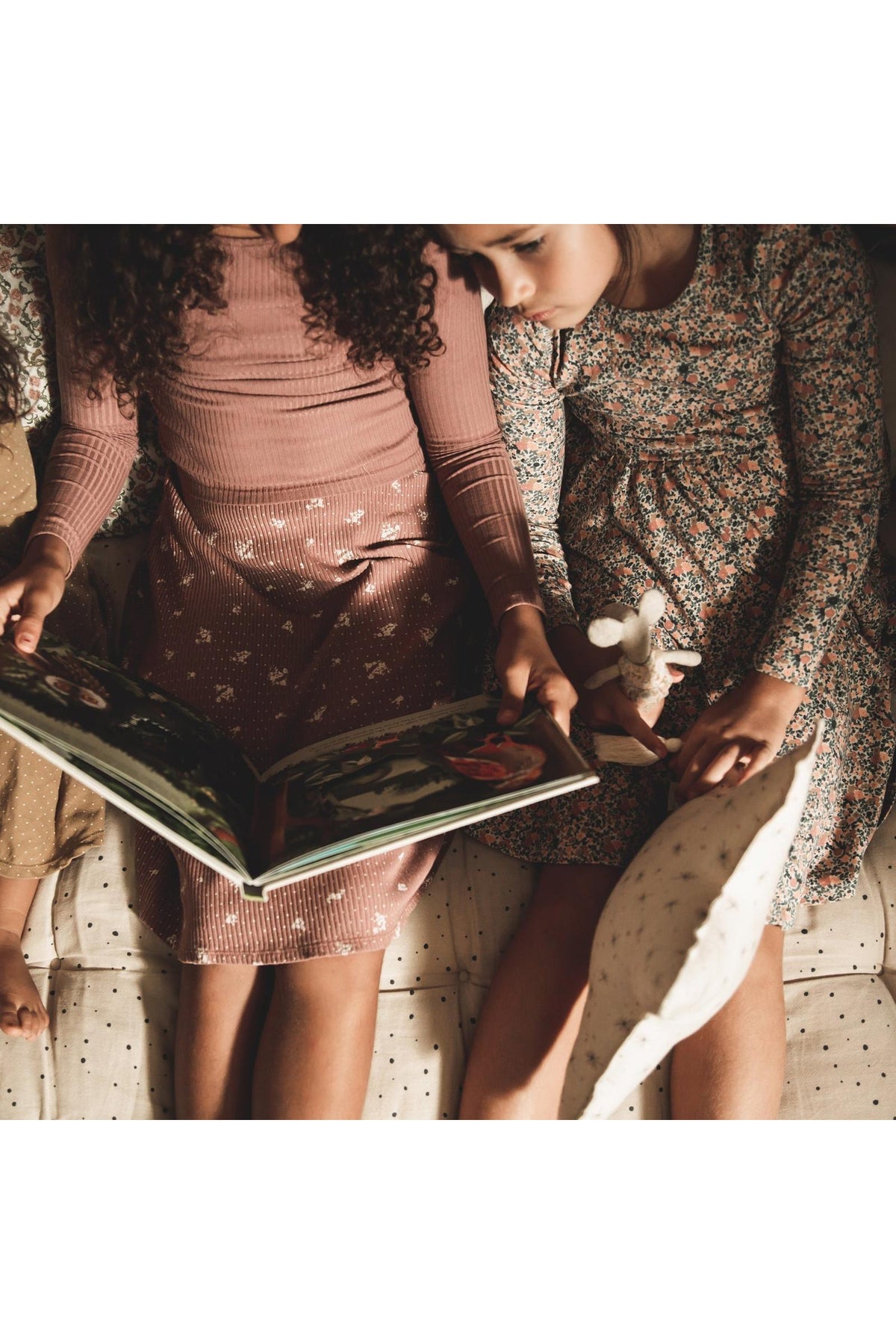 Two children sit closely together, reading a book. One girl wears a rosewood ribbed long-sleeve tee, while the other dons a floral dress, both enjoying a cozy, shared moment.