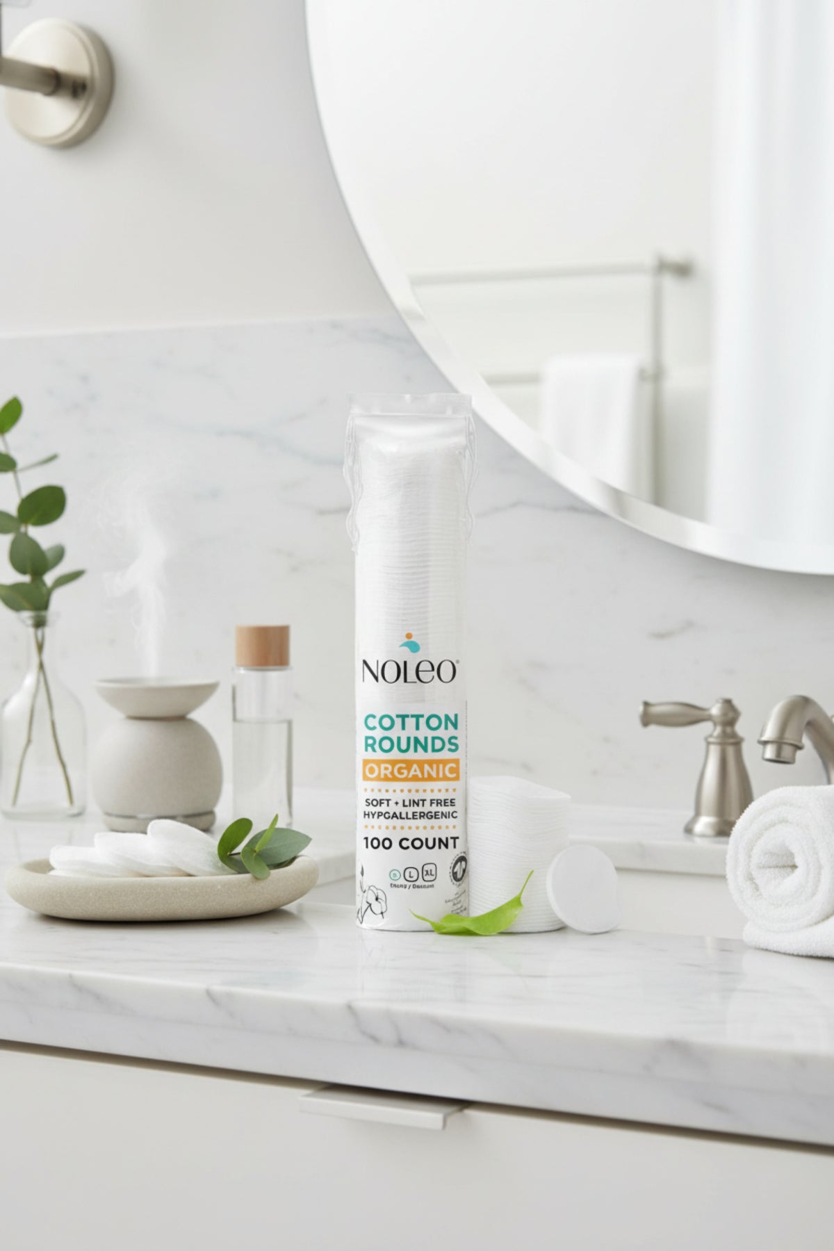 A pack of NOLEO organic cotton rounds is displayed on a marble countertop alongside a small plant and a towel.