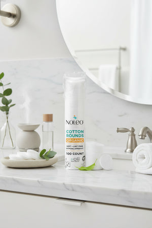 A pack of NOLEO organic cotton rounds is displayed on a marble countertop alongside a small plant and a towel.