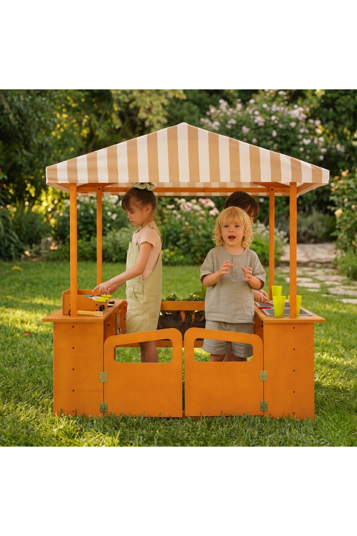 Two children engage playfully at a wooden market stand with a striped canopy, surrounded by lush greenery. The play structure is crafted from sustainable materials, promoting outdoor creativity.