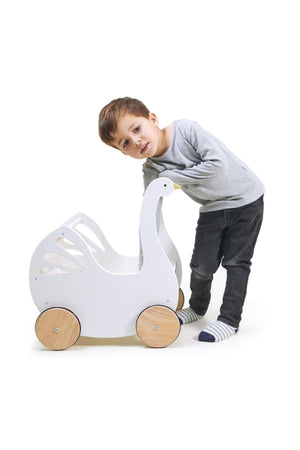 A young boy leans playfully next to a white wooden baby carriage shaped like a swan, featuring natural wooden wheels and cutout details, perfect for imaginative play.