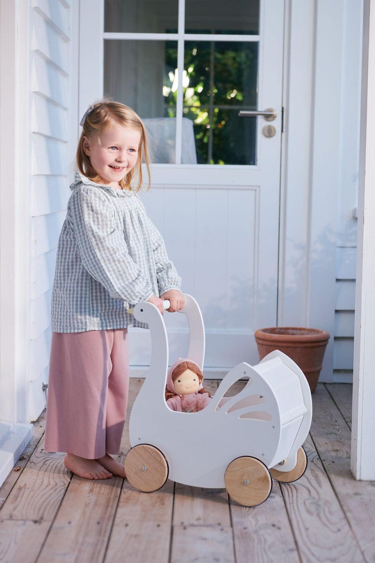A smiling child stands on a wooden porch, pushing a white swan-shaped baby carriage with natural wooden wheels. The carriage holds a doll and features delicate cutout details.