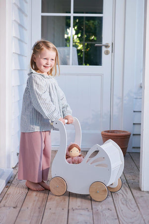 A smiling child stands on a wooden porch, pushing a white swan-shaped baby carriage with natural wooden wheels. The carriage holds a doll and features delicate cutout details.