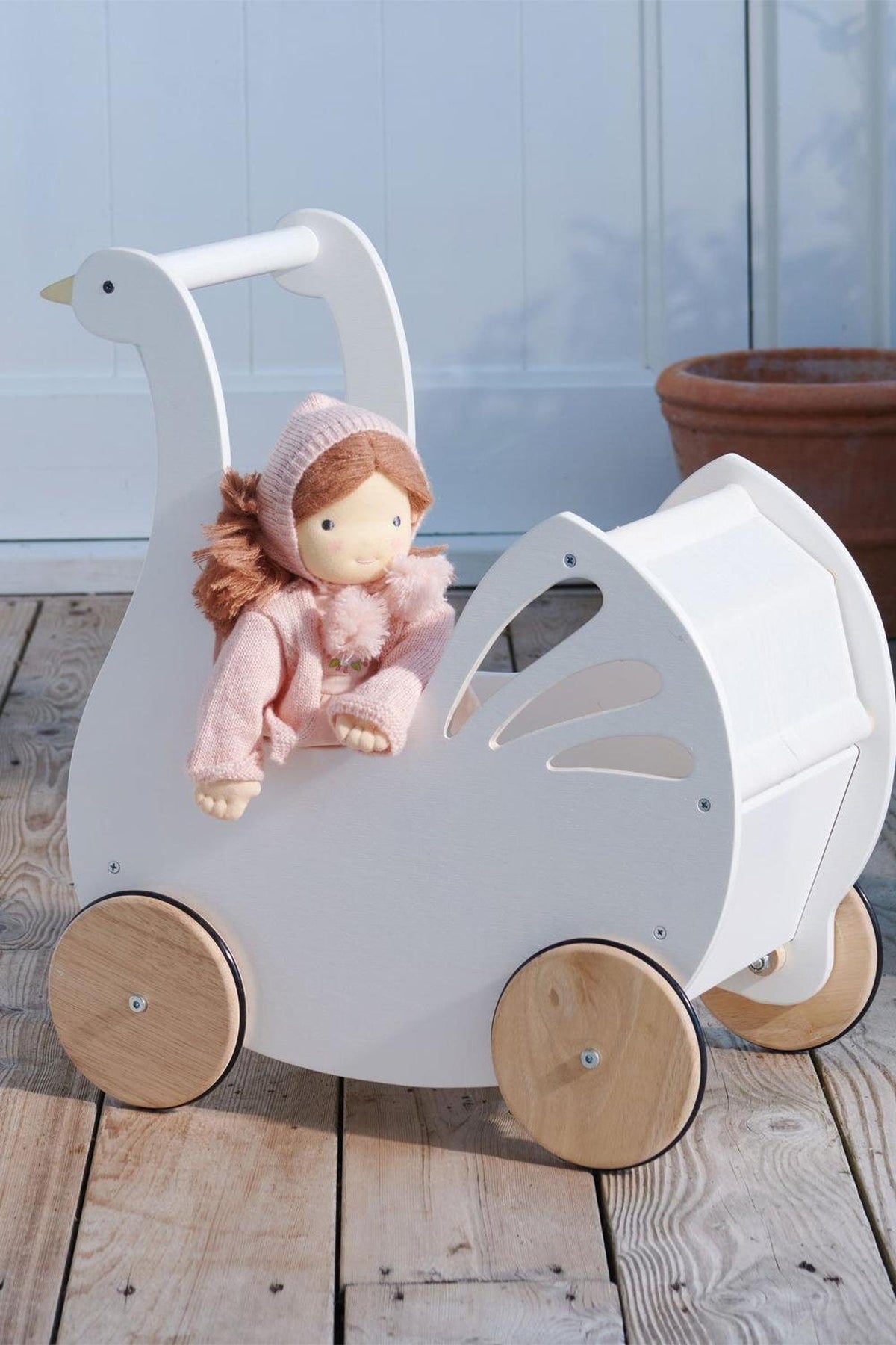 A white wooden baby carriage shaped like a swan sits on a wooden floor, featuring natural wooden wheels. A doll dressed in a pink outfit is comfortably nestled inside.
